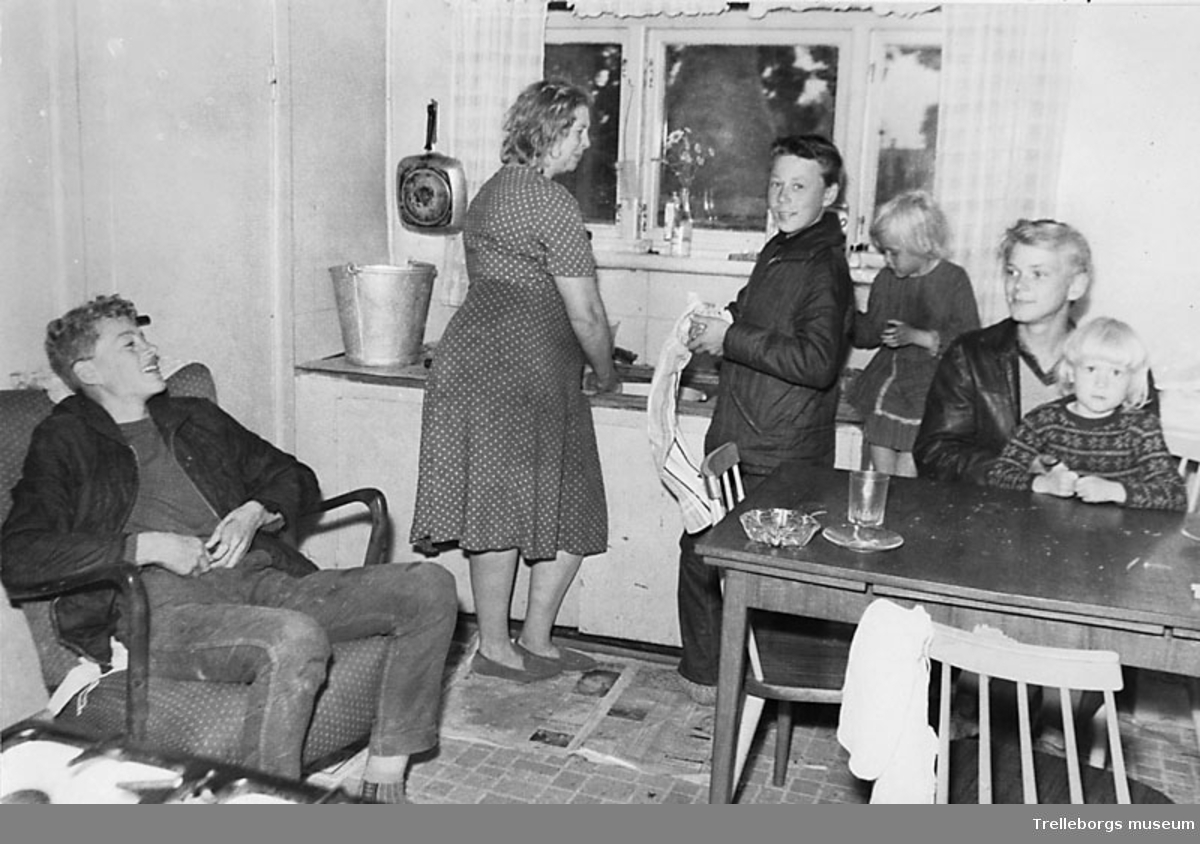 En familj i Fuglie år 1965. Mor diskar och sonen torkar disken. På golvet ligger det tidningar.