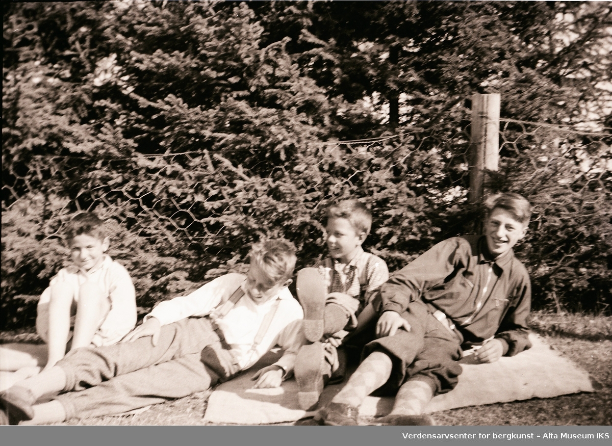 Brødrene Arne, Per og Roar Jøraholmen slapper av på gresset en solfylt dag. Søskenebarnet deres er til høyre. Bildet er fra Jøraholmen, rundt 1937.