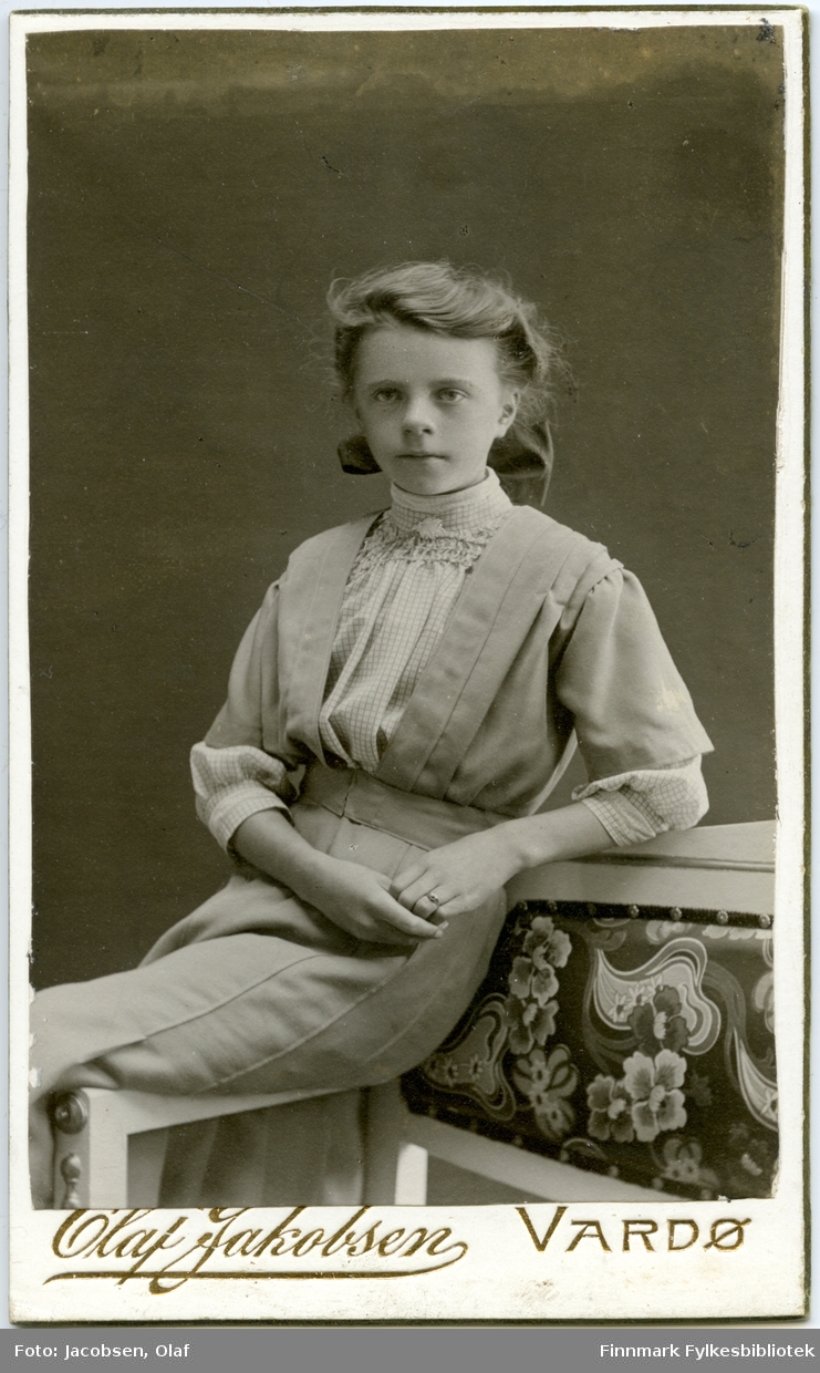 Visitkort. Portrett fra en ung kvinne. Personen ukjent. Hun sitter på armlenen av en sofa og er kledd i en lys kjole.