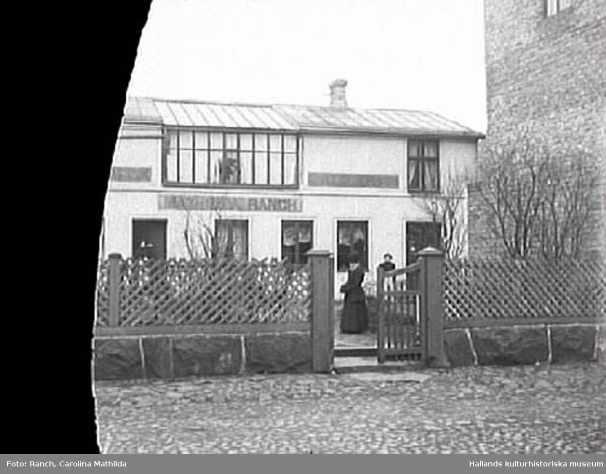 Mathilda Ranchs fotoateljé. Huset låg utmed Prästgatan, kv Bagaren, Varberg. Damen i grinden är Mathilda Ranch. (Glasplåten skadad i vänster sida)
