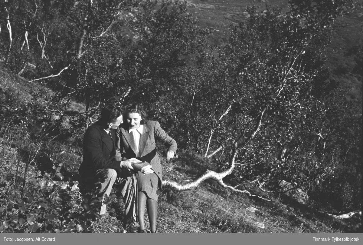 Ragnvald og Alfhild Marie Jacobsen i Jansvannskogen utenfor Hammerfest, sommeren 1947. Alfhild sitter på en grein. Hun har en ganske lyst skjørt på seg og jakke i samme farge. Under har hun en hvit bluse med store snipper og på beina har hun mørke sko. Ragnvald sitter på samme greina. Han har lys bukse og mørk dressjakke. I halsen ses en liten flik av den lyse skjorta og et mørkt, stripete slips. Bakken skrår ganske mye der de sitter og det er endel gress i området. Mye løvtrær og busker i terrenget rundt dem. Sola skinner og det ser ut til å være en flott sommerdag.