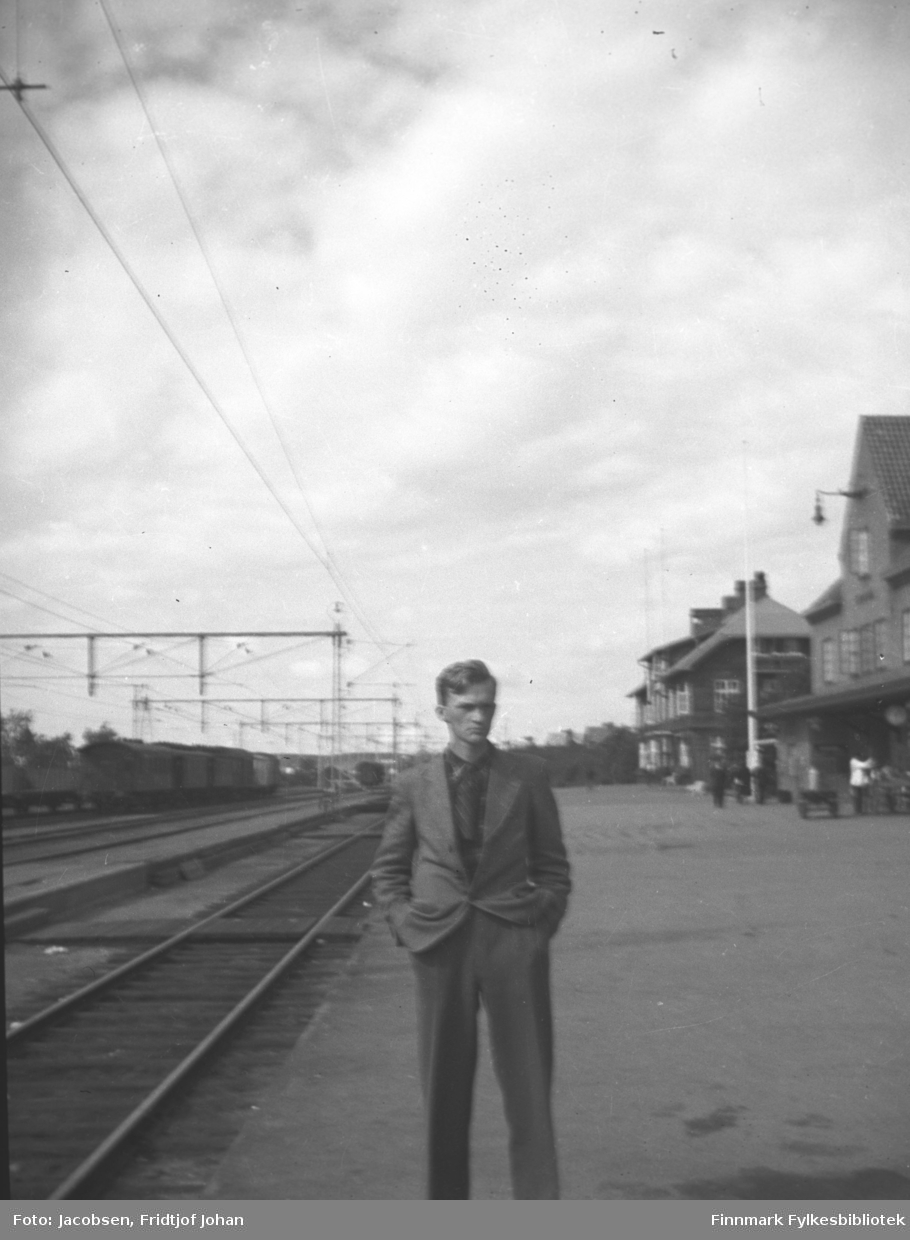 Fridtjof Johan Jacobsen står på en togstasjon/perrong antakelig et sted i Nord-Sverige. Han har en ganske mørk dress på seg. Togskinner og el-linjer samt et tog ses til venstre på bildet. Til høyre står to store bygninger, en stolpe og noen mennesker.