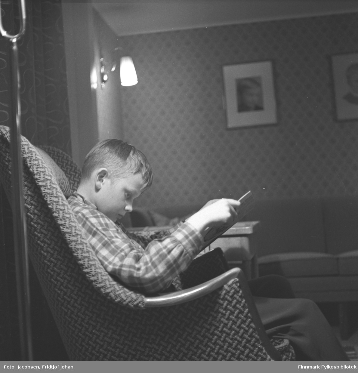 Arne Jacobsen sitter i en lenestol og leser en bok. Et bord står ved siden av lenestolen. Han har en rutet skjorte og ganske mørk bukse på seg. På veggen i bakgrunnen henger en lampe som er tent og et par bilder.