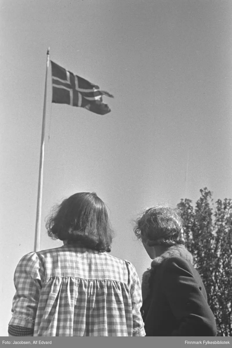 Aase og Ingeborg Jacobsen stående med ryggen til fotografen. De ser opp på det norske flagg som henger i toppen av en flaggstang. Aase, til venstre på bildet, har en rutet sommerkjole på seg. Ingeborg er iført en mørk kåpe/jakke. Helt til høyre på bildet ses et tre med mye løv på. Det er blå himmel og flagget vaier litt i vinden. Iflg. giver av bildet kan det være tatt i mai 1945 på Lødingen.
