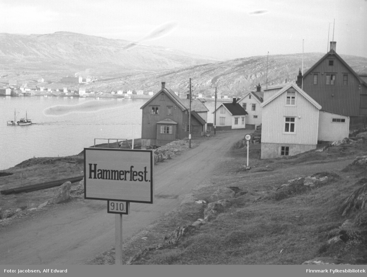 Øvre Haugen, innkjøring til Hammerfest. Et skilt med talltet 910 under bynavnet. Husene står langs veien, som i dag er Rv 94, Akkarfjordveien. Veien som går opp til høyre på bildet er dagens Krutthusgate. I terrenget er det mest gress, lyng og stein. El-stolper står langs veien. Et ganske lite, lysmalt hus med ark nærmest kamera til høyre på bildet. Det har tre vinduer på røstveggen og stående panel. Et større hus står ved siden av det. Huset er ganske mørkt med lys grunnmur. Flere vinduer ses på røstveggen. Et fartsgrenseskilt står i veikrysset. Huset nærmest på venstresiden av veien er forholdsvis lyst med stående panel. Det har små tilbygg, både på røstveggen nærmest og på andre siden. Flere hus står nedenfor dette, alle er lyse/hvite. Det lille i midten har mørkt takdekke, mens de to andre har skifer. En stor skøyte er på vei ut av havna. Over havna ses deler av bydelen Fuglenes. Hus står langs Fuglenesveien og noen større bygg helt til venstre på bildet. Det store bygget litt ovenfor strandkanten/Fuglenesveien, er sykehuset. Fjellet bak sykehuset er Storfjellet og til høyre ligger Fuglenesfjell. Det ser ut til å være en værmessig fin sommerdag.