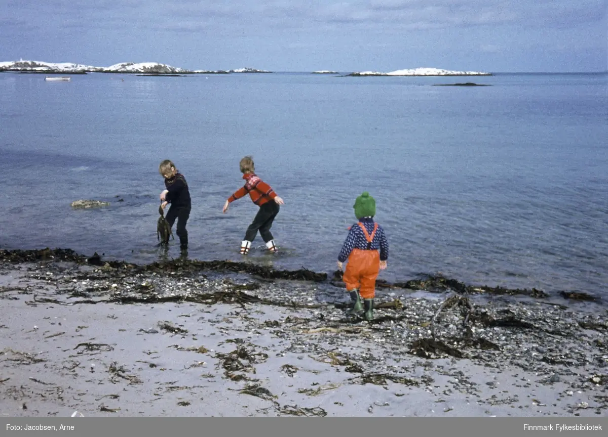 Tre barn leker i fjæra på Andenes. Fra venstre: Mona Stenmann, Elisabeth Vråli og Tor Jørgen Jacobsen. Tor Jørgen har regnbukse med sele, gummistøvler, lue med dusk og mønstret ullgenser. De to jentene, som vasser, har også gummistøvler, mørke busker og ullgenser. Elisabeth kaster stein i sjøen. Det er havblikk og snø ligger på holmene og neset i bakgrunnen. Toppen av en veske/bag kan ses i nedre billedkant.