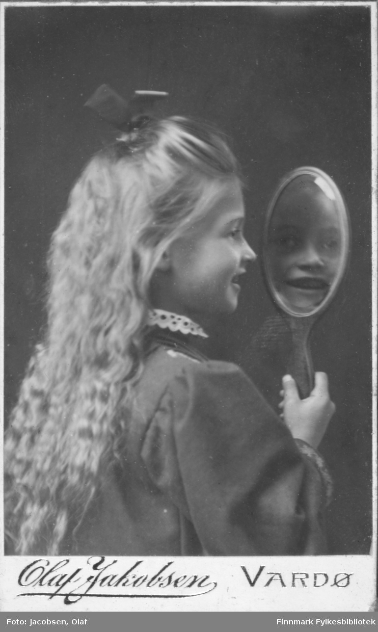 Portrett av en ukjent jente som holder et speil. Jenta står på siden og ser seg i speilet. Hun har langt hår som er pyntet med en hårsløyfe. Rundt halsen på kjolen eller jakken har hun en blondekrage