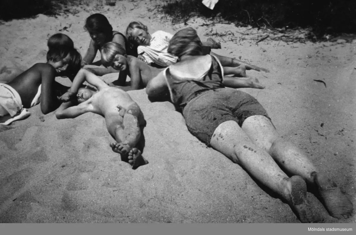 Två kvinnor och fyra barn ligger och solar på sanden, troligtvis Tulebostranden cirka 1930.