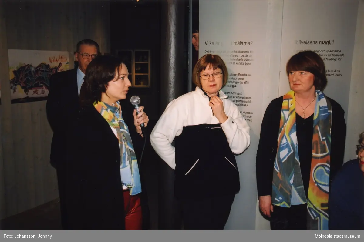 Invigning av utställningen "Alltså finns jag" på Mölndals museum år 2003. Visning av utställningen skedde av konstpedagog Karin Lundmark från Bohusläns museum, som här håller i mikrofonen, samt arkeolog/pedagog Anna Bratås från Vitlycke museum (ej i bild). Till höger ses museichef Mari-Louise Olsson. En okänd man skymtas i bakgrunden till vänster.