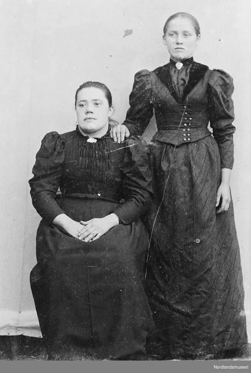 Fagfoto av 2 kvinner i helfigur. Kvinnen til venstre sitter mens den andre står. De  er begge kledd i mørke lange kjoler. I halsen på kjolene er det festet en nål.