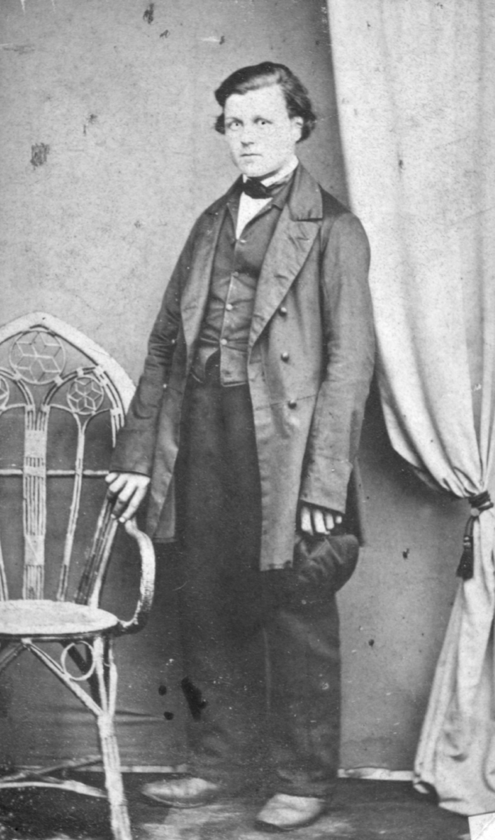 Portrett av Henrik Jonas som emmigrerte til Seattle, Amerika, USA. Flere i hans slekt emigrerte til Amerika. Han er kledd i jakke, vest, bukser og sko. I sin venstre hånd holder han på en hatt. Han hviler den høyre armen sin på en stol. Bak ham kan man se en gardin.