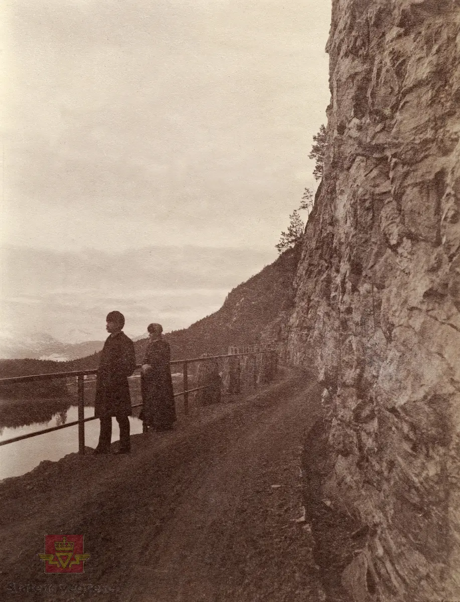 "Veianlæg". Dame og mann stående på veg med rekkverk av jern, stabbesteiner og bratte fjell. Ukjent sted ca. 1924.