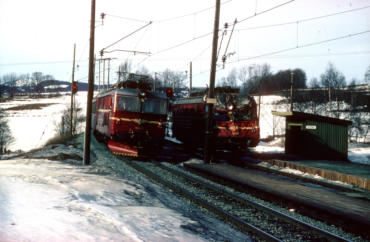 Ekspresstoget Dovreekspressen, tog 42, kjører forbi El 14 2197 på Nypan stasjon. 2197 er satt på traller etter kollisjon og skal fraktes til Grorud.