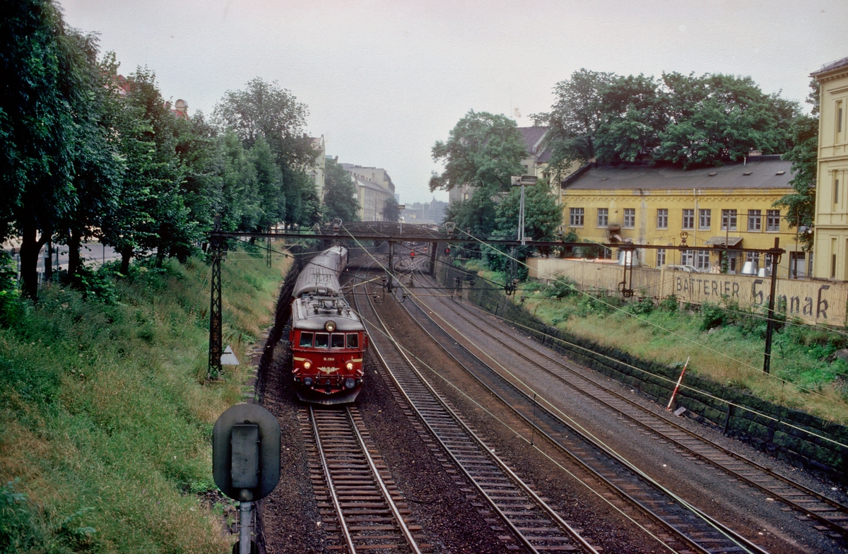 NSB persontog til Vestfoldbanen kjører ut av "Strupen" fra Oslo V. Lokomotiv El 11 2108. Bildet er tatt fra Munkedamsveiens bro over Drammenbanen, og forbindelsessporene til Filipstad. Reklame fra Sønnak på gjerdet til høyre.