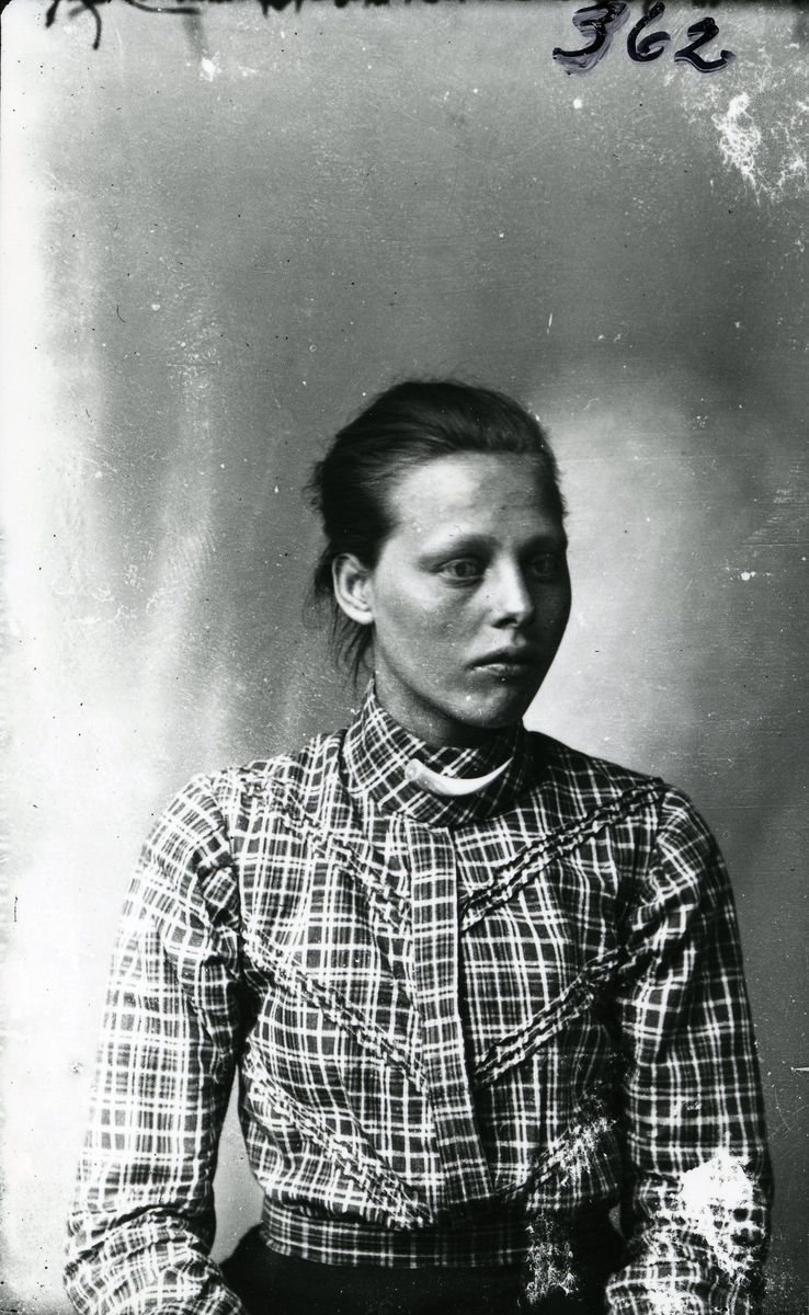 Portrett i halvfigur av kvinne sittende foran lerret. Rutete bluse og svart skjørt.