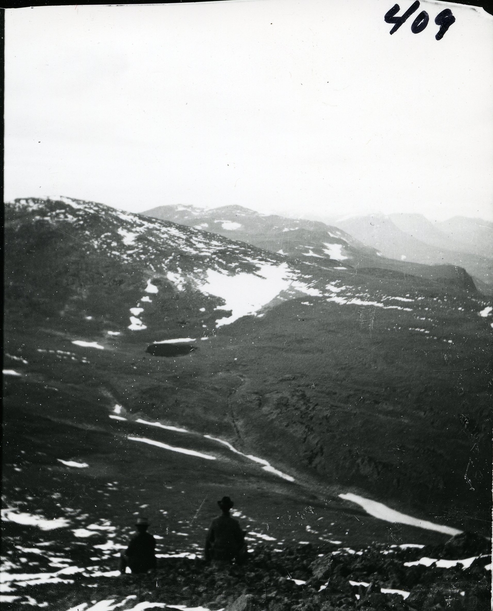 To menn sitter og skuer ut over fjellheimen. Begge med hatt. Foto tatt med Skogshorn (Hemsedal) i ryggen og med utsyn over Vangsfjella. Ranastonge, Rankonøse og Grindadn lengst bort.