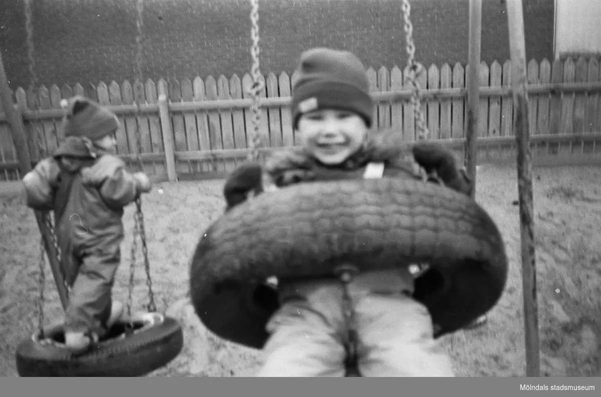 Två små barn gungar på var sin gunga. Ställningen står i en stor, inhängnad sandlåda utanför Katrinebergs daghem, 1992.