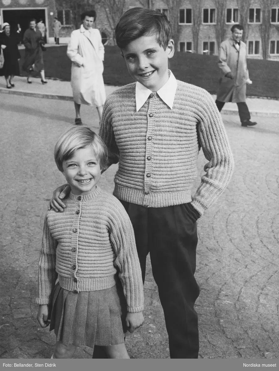 Två barn i likadana stickade koftor, flicka i kjol och knästrumpor, pojke i byxor.