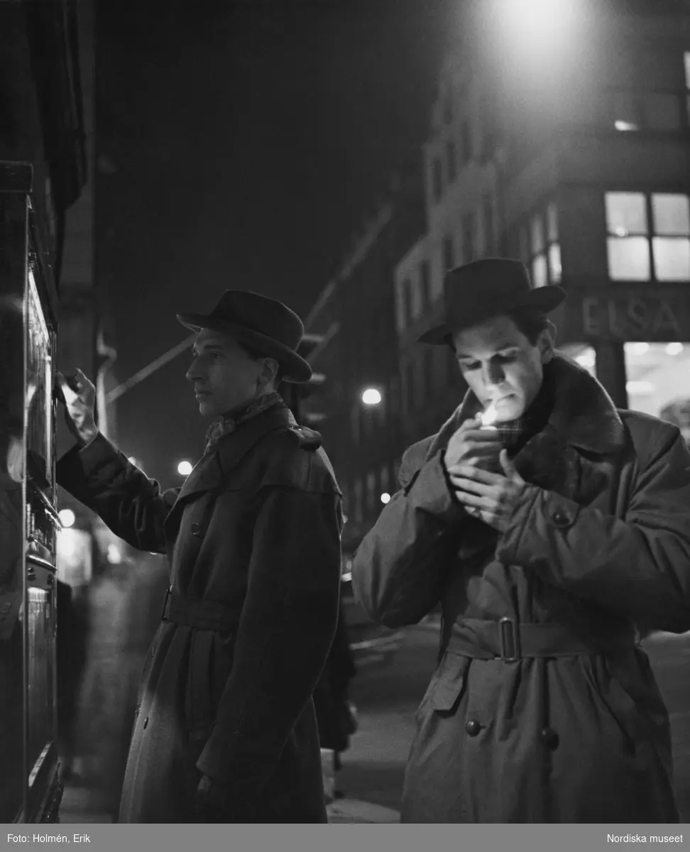 Två män vid tobaksautomaten utanför Nordiska Kompaniet. Båda är klädda i trenchcoat och hatt, den ene tänder en cigarrett.