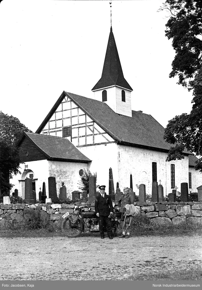 Borre kirke. Mann i uniform og dame stående foran kirken med motorsykkel.