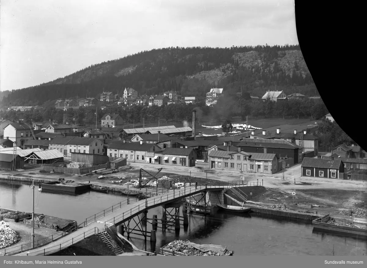 Vy över Norrmalm. Gamla gångbron Puckelbron, även kallad "Kamelryggen" över Selångersån (låg i Norrlandsgatans förlängning, raserades 1919 vid översvämningen). Den ersattes med en ny bro (som också kallades Puckelbron) i Malmskillnadsgatans förlängning. Glasplåten är skadad (en bit saknas i övre högra hörnet).