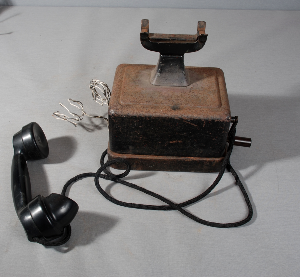 Telefonapparat med telelfonrør i gaffel på toppen av apparatet, sveiv på siden. Selve kassen står på sokkel av metall. 2 ledninger festet på siden av apparatet.