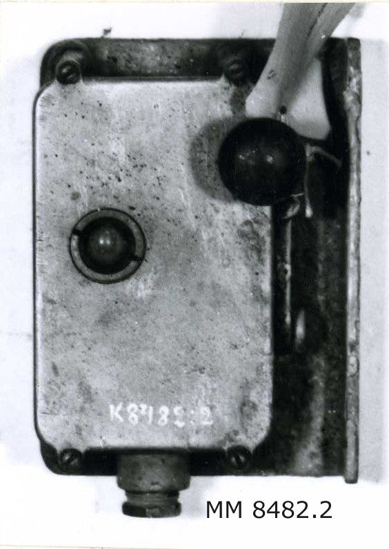 Signalapparat med telegrafnyckel i kåpa av lättmetall med kabelintag och öron för fastsättning. Märkning: 4475.
Fastsatt på en järnplåt med uppvikt kant.