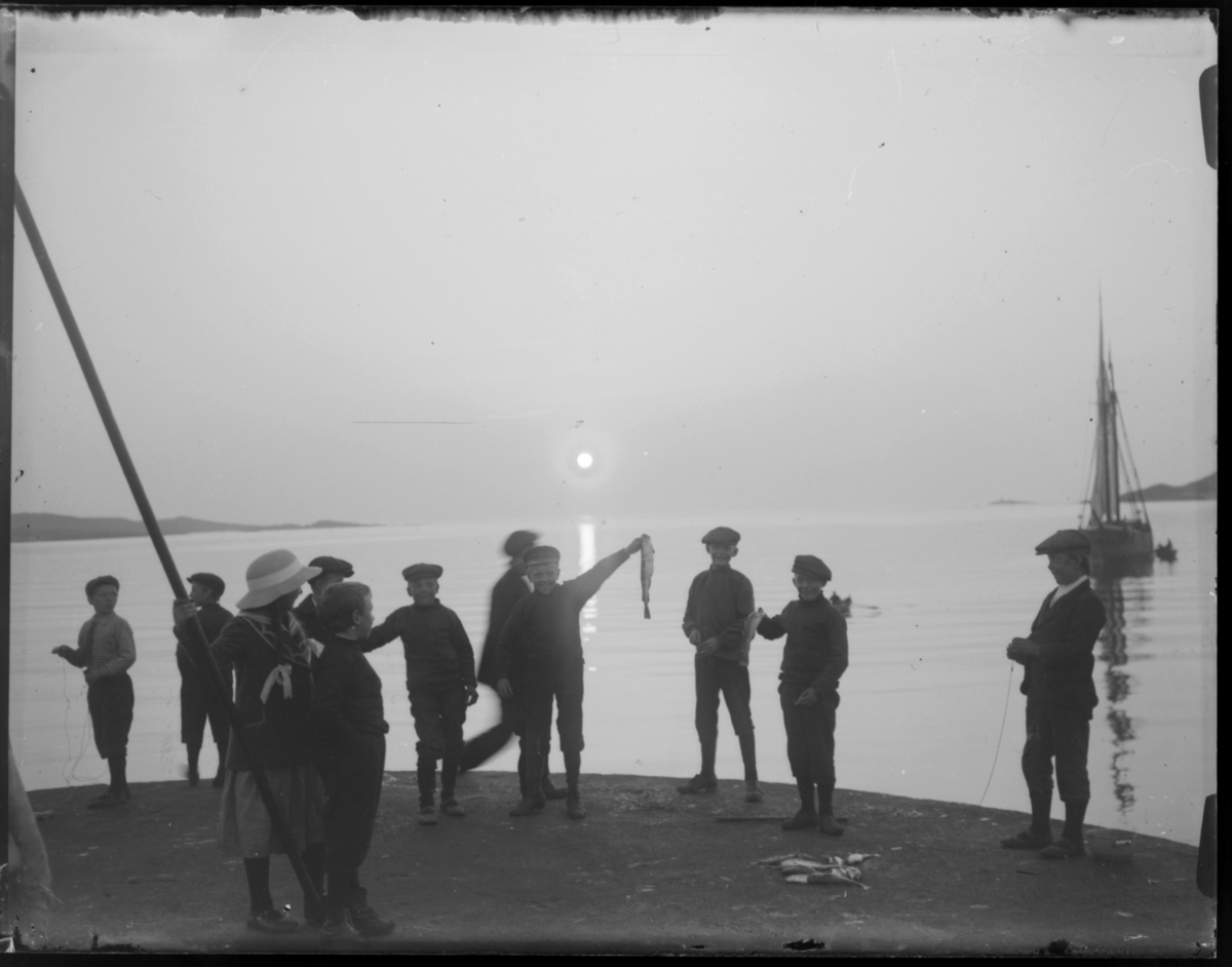 Noen gutter står ytterst på østre molo i Vardø i midnattsola og fisker med snøre. De har fått noen småfisk som de stolt viser fram til fotografen. I bakgrunnen ser man Skagen til venstre og Reinøya til høyre