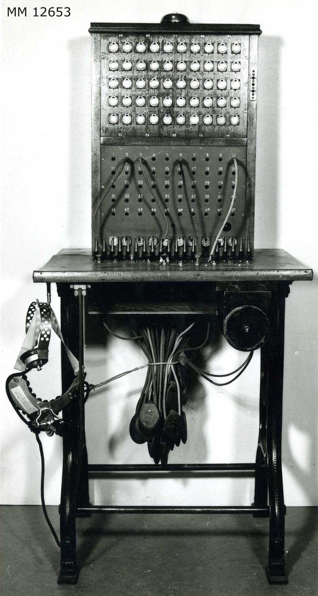 Telefonväxel typ OB, snörväxel, 50 linjers. Försedd med hand- och bröstmikrotelefon. Växelbordet med panelen vilar på järnstativ.