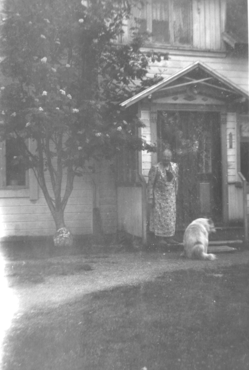 En dame med en lys hund står foran innganegen til et hus. Ved siden av inngangen et stort tre.