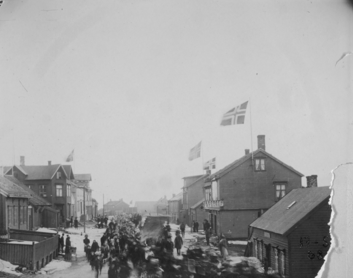 Folk som går i tog. Flagg på husene. Bildet er tatt fra Kr. IVs gt. matr. 35 mot Østervågen og resten av gaten.