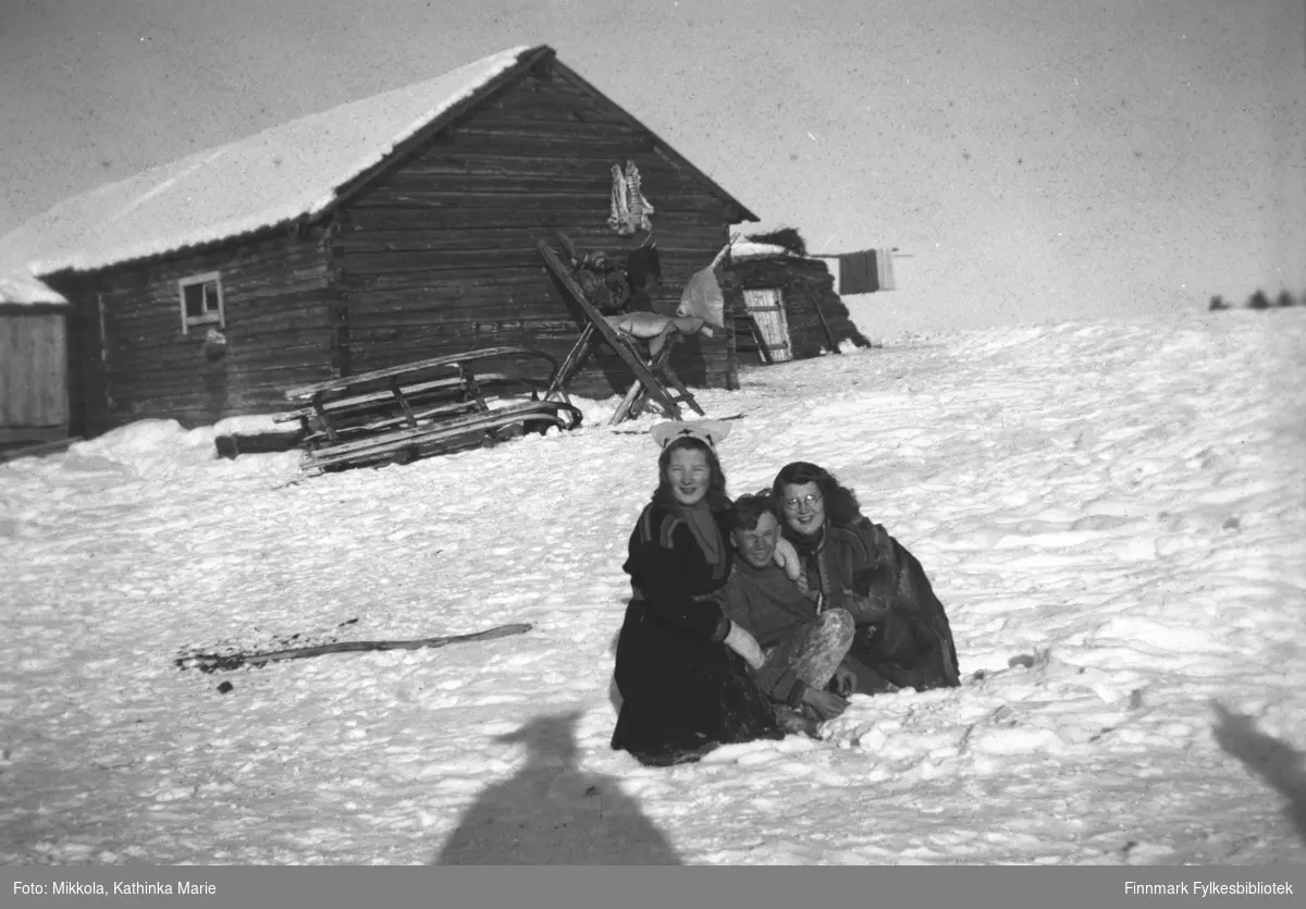 Pakanajoki, ca. 1946-1947. Fra venstre: Kari Mikkola, Valle med ukjent fornavn, Ingrid Mikkola. I bakgrunnen en tømmerhytte med diverse utstyr lagret utenfor. Vi ser bl.a. en hvelvet slede og et lagringsstativ av tre. Til høyre for hytta står en gamme