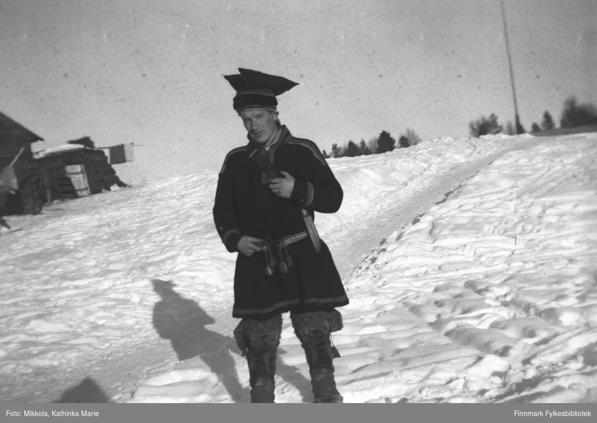 Andreas Mikkola i kofte, bellinger og med stjernelue og kniv i beltet. Bildet er tatt i Pakanajoki ca. 1946-1947. I bakgrunnen til venstre en gamme