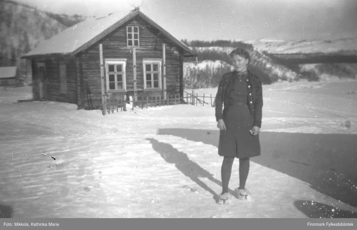Gudrun Mikkola fotografert i snøen foran gamlestua på eiendommen Nordmannseng. Eiendommen ligger ved kirka i Neiden. Denne tømmerstua ble også brukt som skole i noen perioder.
