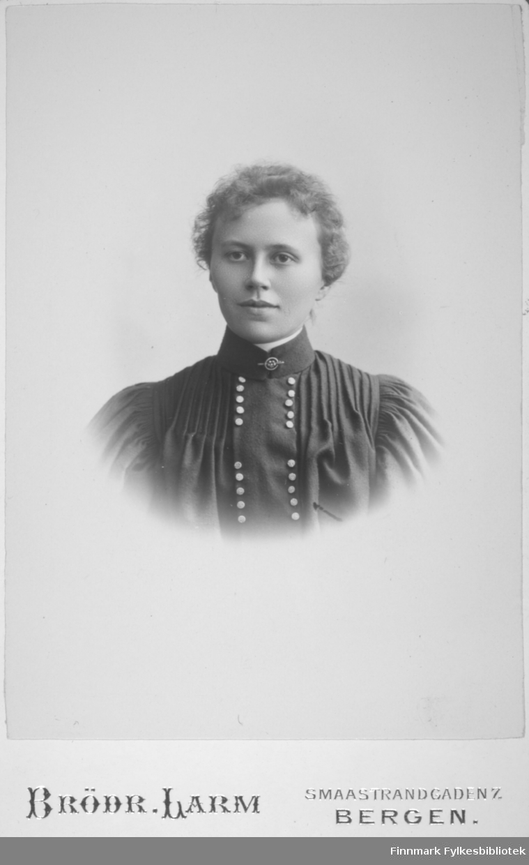 Portrett av en dame i mørk kjole. Lyse knapper går tett i tett fra halsen og nedover brystet. Portrettet er tatt hos Brødrene Larm i Bergen.