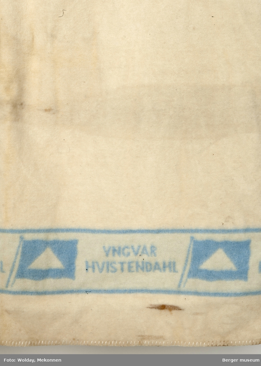 Hvitt teppe med logo for Yngvar Hvistendahls rederi på lysblå stripe i hver ende av teppet. Tekstfelt invertert på andre siden av teppe
