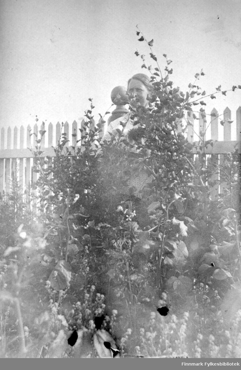 En dame står delvis skjult bak en høy plante i Arthur og Kirsten Bucks hage. Damen har en hvit overdel på seg. Det er mye planter som står ganske tett. Et hvitt stakittgjerde står rett bak damen. En av stolpene med dreiemønster og kule på toppen ses rett bak hodet hennes.