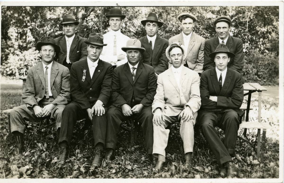 Gruppebilde av ti menn sittende på en benk. Alle kledd i dress og hatt.