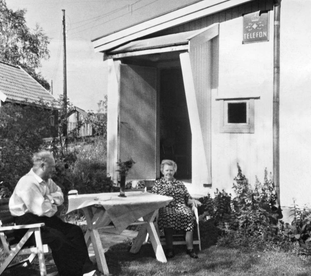 Bjørkelangen telefonsentral, tidlig 1960-tall.
Stasjonsholder Alfred Lunner (tv) og hans kone Mathea ved hagebordet utenfor sentralen. Legg merke til telefonskiltet til høyre på veggen. I et lite rommet innenfor det lille vinduet kunne publikum bestille telefonsamtaler.