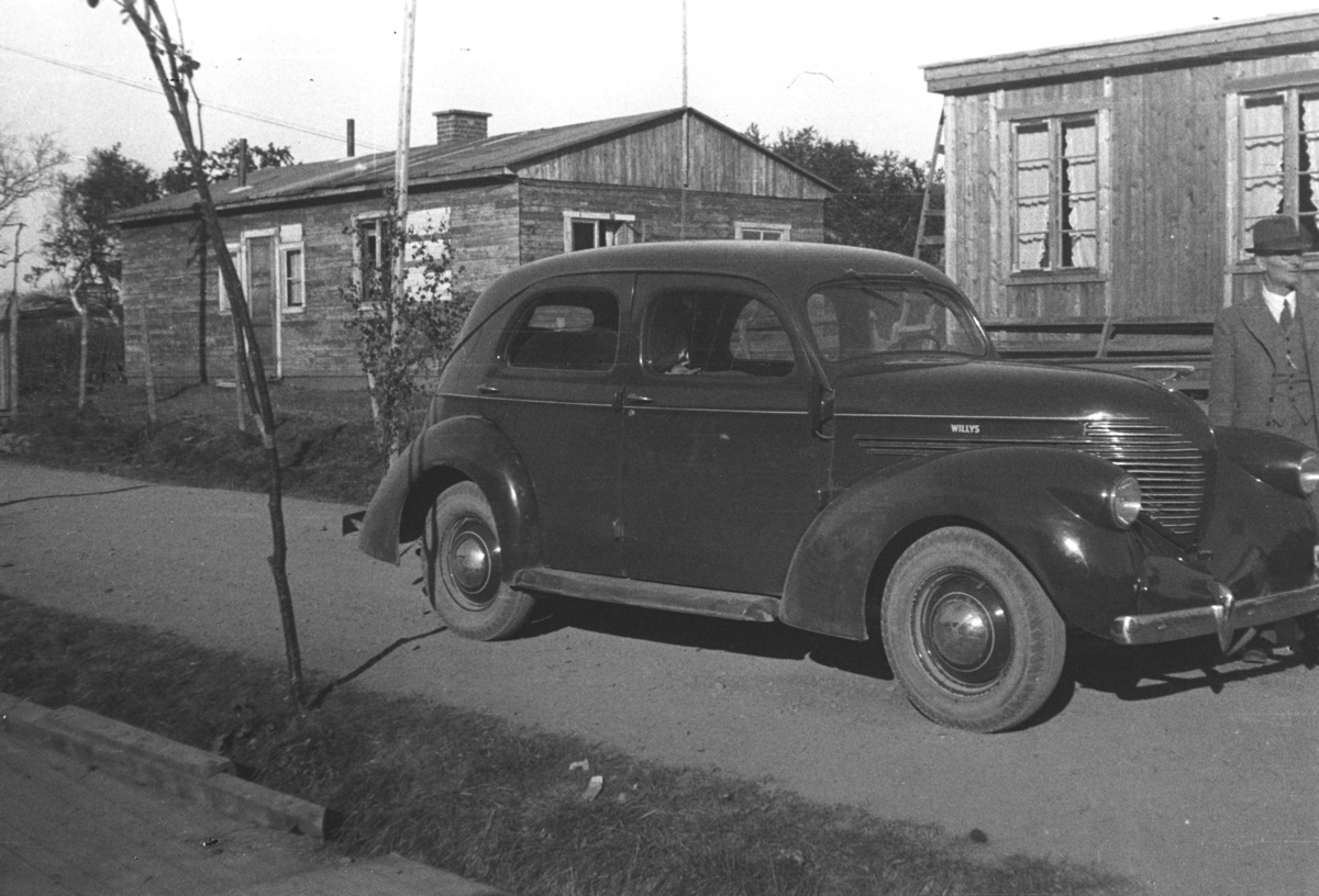 En bil (Willys 1937-38-modell) står parkert utenfor to små hus eller hytter. Stedet er ukjent, men iflg. informanten kan det være i Tana.