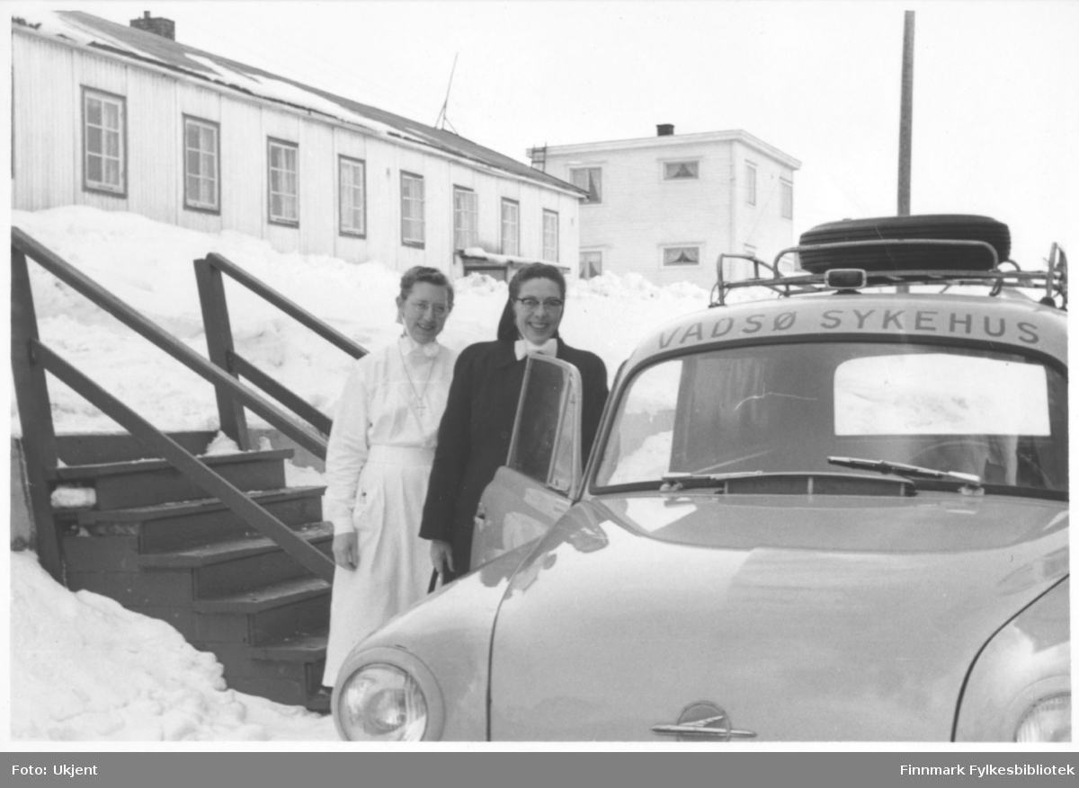 Reisende sykepleier Ingeborg Søvik står ved siden av en bil med tekst Vadsø sykehus (Opel Olympia varebil årsmodell 1953-57). Til venstre for henne står oversykepleier Gudrun Byholt. I bakgrunnen kan man se søsterbrakka. Gudrun har på seg uniform og et kors rundt halsen. Ingeborg har på seg jakke. De har begge briller. På taket på bilden kan man se bagasje. Ved siden av kvinnene kan man se en trapp. Det er snø på bakken. På bygningene kan man se vindu og skorsteiner.