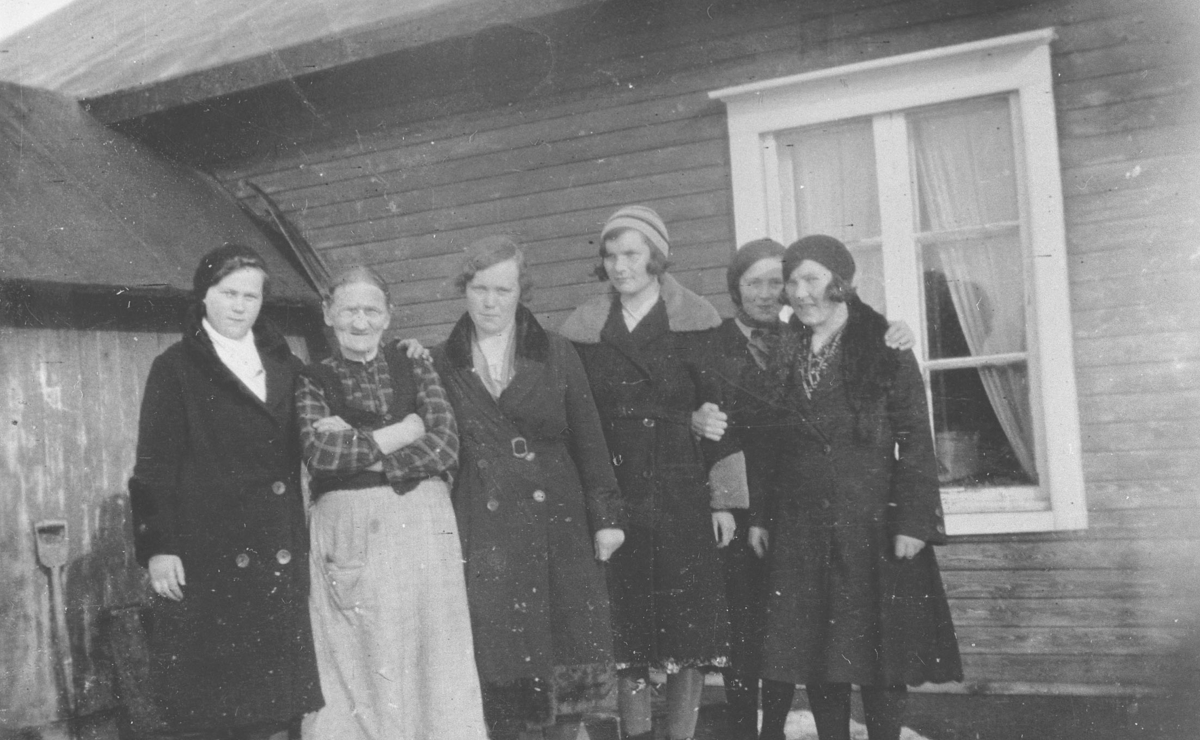 Seks kvinner fotografert ved et hus i Vestre Jakobselv. Kvinnene fra venstre: Emma Reisænen, hennes mor Brita Stina Mietinen (f. Dørmænen i Kuusamo, Finland), Hilda Severine Thuv, Elise Henninen, Miriam Mietinen og Petra Ballo. Kvinnene har vinterkåper på seg. Bildet er antakelig tatt om våren, for det ligger fortsatt litt snø. Bak kvinnene et vindu med lyse gardiner