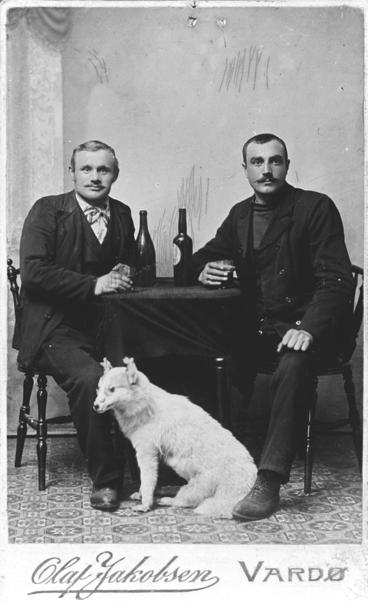 To menn, til venstre Oskar Ittelin, sitter ved bord i atelieret til Olaf Jakobsen. På bordet står to vinflasker og to glass. Bordet er dekket av en mørk duk. Mennene holder på glassene sine og ser rett på kamera. Foran dem på gulvet sitter en utstoppet rev eller et lignende dyr
