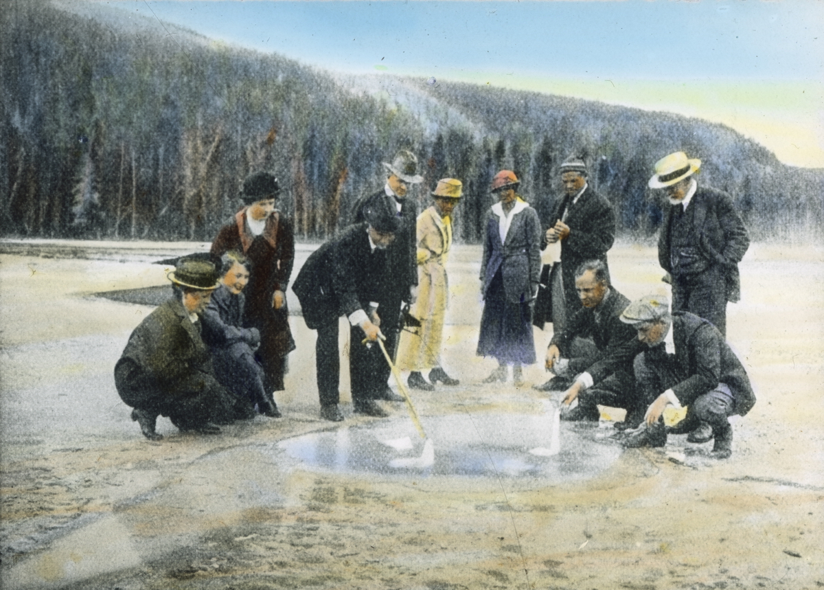 Håndkolorert dias. Avfotografert fra ukjent original. En gruppe kvinner og menn står og sitter rundt en geysir som bobler.