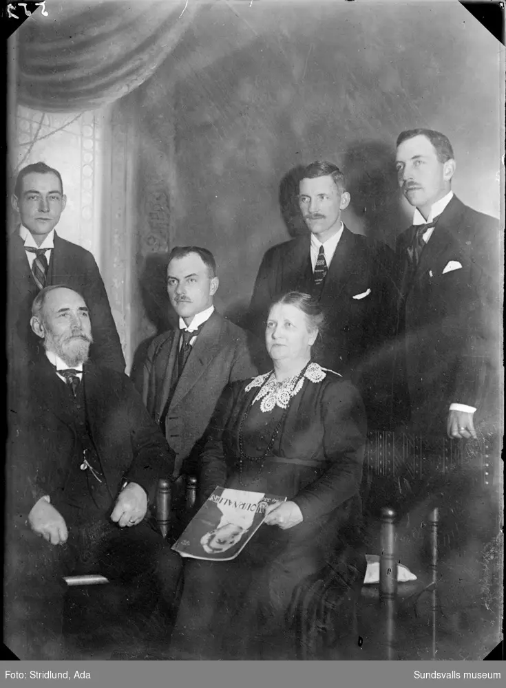 Fem män och en kvinna. Kvinnan sitter i en stol och håller en tidning i handen. Ateljéporträtt.