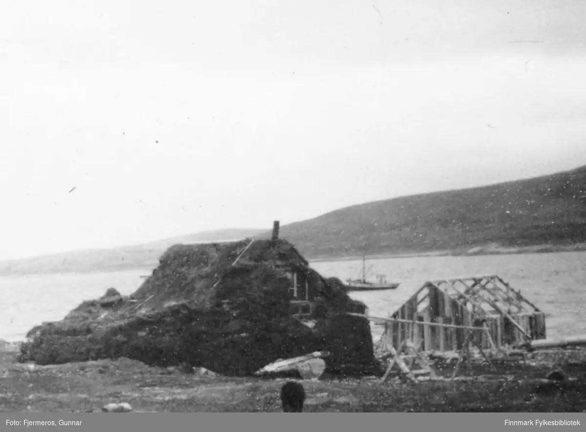 Et lite hus dekket av torv, sansynligvis for isolering. Et lite bygg som kan være et naust/sjøbod, er under oppføring like ved. En sjark ligger fortøyd ute på sjøen. Stedet er ukjent. Bildet er tatt våren 1946.