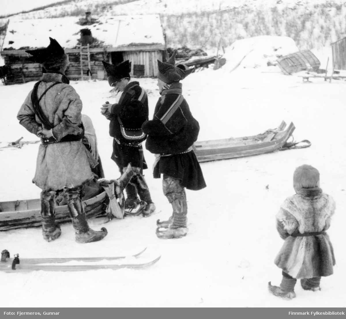 Fotografi av tre voksne og et barn i sameklær med noen sleder og ski på den snødekte bakken. Bildet er tatt på Jergul i påsken 1947.