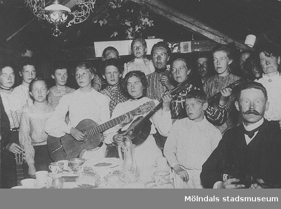 Män, kvinnor och barn står vid ett kaffebord, några av dem håller i instrument. Okända namn, plats och årtal.