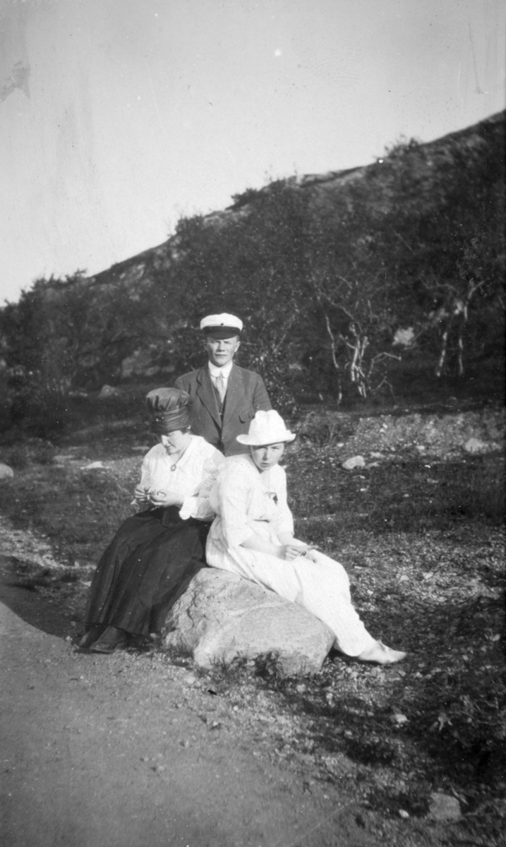 "Fra Soldatbukten. Knes." To kvinner sitter på en stein ved siden av en grusvei. De er kledd i kjoler. En ung mann står bak dem, og har en hvit hatt på hodet. I bakgrunnen kan man se skog.
