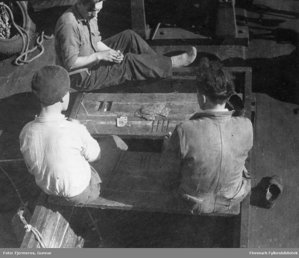 Tre unge menn sitter på dekket til en fiskeskøyte og spiller kort. Menn og båt er ukjent, men bildet er tatt i Laksefjorden våren 1948.